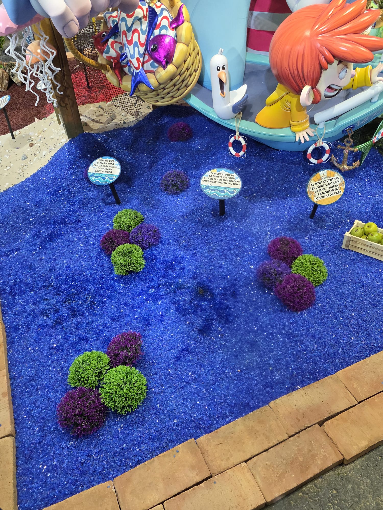 Detalle de la decoración de la hoguera infantil Mercado Central 2026, donde el cristal triturado azul recrea el agua y aporta luminosidad a la escena.